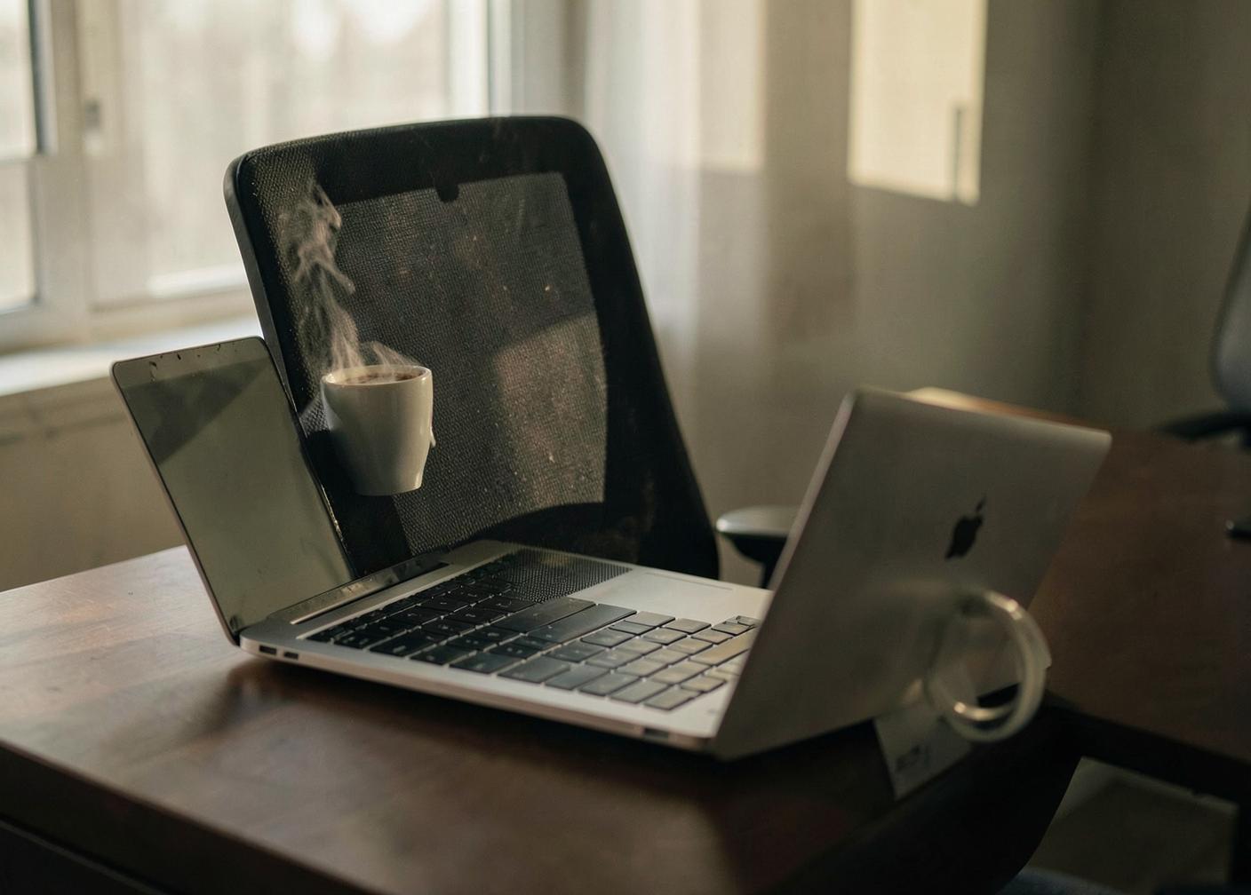 An empty office chair pushed back from a desk with a laptop left open and a cold cup of coffee